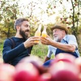 Zwei Männer sitzen vor Apfelbäumen und stoßen mit Apfelsaft an.