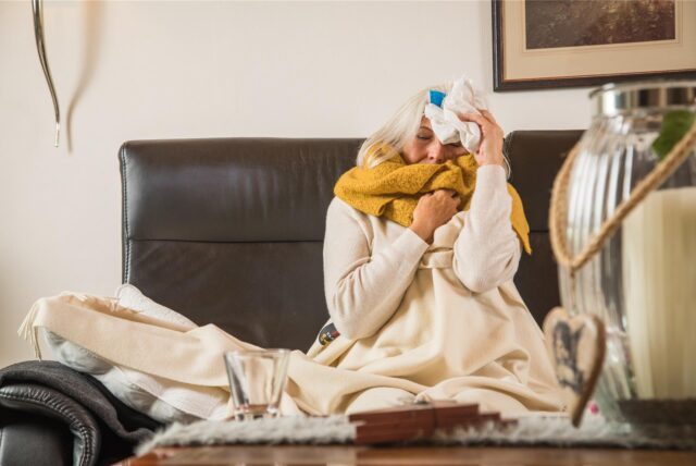 Eine Frau liegt mit Decke und Schal auf dem Sofa und hält sich ihren Kopf.
