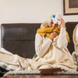 Eine Frau liegt mit Decke und Schal auf dem Sofa und hält sich ihren Kopf.