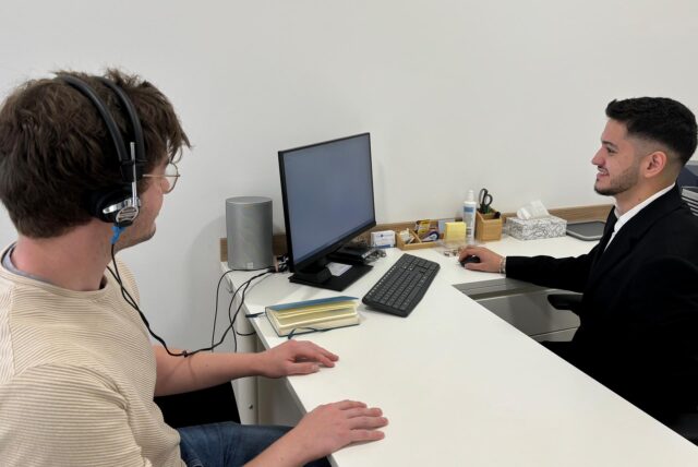 Kerim Aydin sitzt rechts im Bild an einem Tisch und bedient einen Computer. Der Tisch und die Wände um ihn herum sind alle weiß. Ein paar Bürounterlagen liegen auf dem Tisch. Auf der anderen Seite sitzt ein Mann mit Kopfhörern. Sie sind mit dem Computer verbunden.