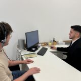 Kerim Aydin sitzt rechts im Bild an einem Tisch und bedient einen Computer. Der Tisch und die Wände um ihn herum sind alle weiß. Ein paar Bürounterlagen liegen auf dem Tisch. Auf der anderen Seite sitzt ein Mann mit Kopfhörern. Sie sind mit dem Computer verbunden.