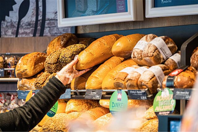 Eine Verkäuferin greift mit einem Einmalhandschuh nach einem Brot in einem Verkaufsregal. Verschiedene Brotsorten sind in dem Regal ausgelegt und mit Preisschildern versehen.