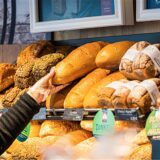 Eine Verkäuferin greift mit einem Einmalhandschuh nach einem Brot in einem Verkaufsregal. Verschiedene Brotsorten sind in dem Regal ausgelegt und mit Preisschildern versehen.