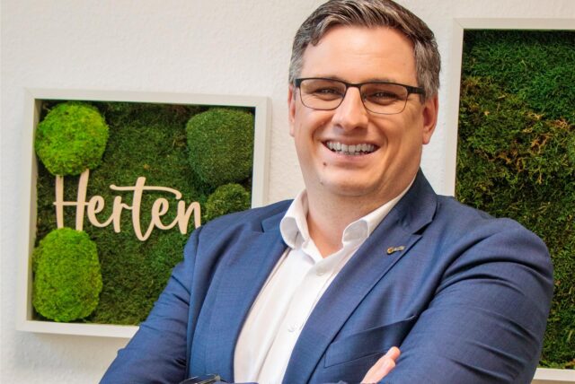 Patrick Berner lacht in die Kamera; hinter ihm gerahmte Fotos mit dem Schriftzug Herten an weißer Wand.