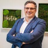 Patrick Berner lacht in die Kamera; hinter ihm gerahmte Fotos mit dem Schriftzug Herten an weißer Wand.