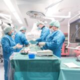 Vier Ärzte und Pflegepersonal in blauen Kitteln an einem OP-Tisch in einem Krankenhaus.