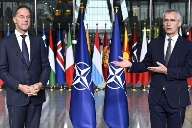 Mark Rutte steht links im Bild, rechts Jens Stoltenberg und spricht in ein Mikrofon. Zwischen ihnen stehen NATO-Flaggen.