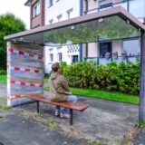 RZ-Redakteur Tobias Mühlenschulte sitzt mit dem Rücken zur Kamera auf einer Holzbank in einem Fahrgastunterstand der Vestischen Straßenbahnen an der Bushaltestelle Röllinghausen Kirche an der Winnlohstraße. Die rechte Seitenscheibe und die hintere Scheibe des Wartehäuschens fehlen. Im Hintergrund ist ein Riegel aus Wohnhäusern zu erkennen – teilweise mit Balkonen.