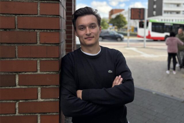 Julian Schallkamp steht in der Hertener Innenstadt und lehnt an den  Pavillon am Eingang der Herrmannstraße. Im Hintergrund stehen Fußgänger und im Kreisverkehr fährt ein Bus und ein Auto.