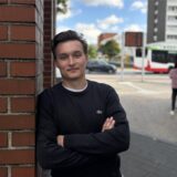 Julian Schallkamp steht in der Hertener Innenstadt und lehnt an den  Pavillon am Eingang der Herrmannstraße. Im Hintergrund stehen Fußgänger und im Kreisverkehr fährt ein Bus und ein Auto.