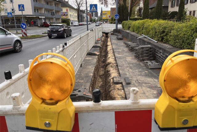 Eine Baustelle: Am Fahrbahnrand sind Baustellenabsperrungen aufgestellt die einen Graben direkt am Rand des Bürgersteiges umzäunen.
