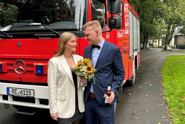 Ein Brautpaar steht vor einem Feuerwehrauto. Sie hält einen Blumenstrauß in der Hand, er ein Bier.