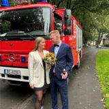 Ein Brautpaar steht vor einem Feuerwehrauto. Sie hält einen Blumenstrauß in der Hand, er ein Bier.
