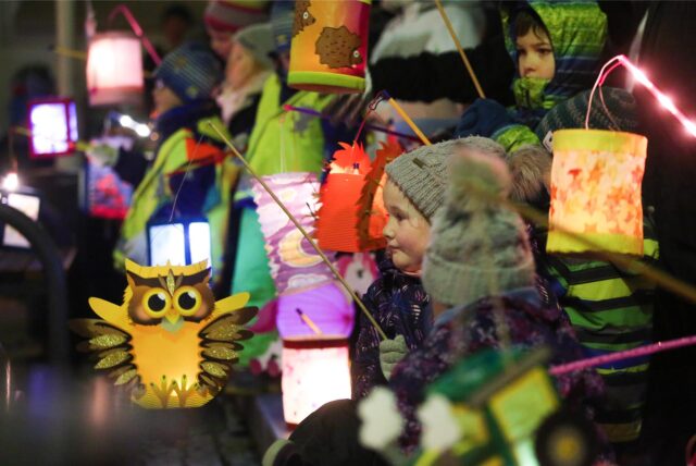 Ein junges Mädchen steht, dick eingepackt in Mütze und Winterjacke, in einer großen Kindergruppe. Sie hält eine leuchtende Laterne, die wie eine Eule aussieht. Die anderen Kinder, halten auch ihre Laternen in der Luft.