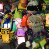 Ein junges Mädchen steht, dick eingepackt in Mütze und Winterjacke, in einer großen Kindergruppe. Sie hält eine leuchtende Laterne, die wie eine Eule aussieht. Die anderen Kinder, halten auch ihre Laternen in der Luft.