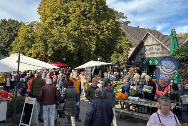 Der Spargelhof Schulte-Scherlebeck ist vom Eingangstor aus zu sehen. Es sind viele Sonnenschirme aufgestellt und eine Menschenmenge ist läuft umher. Tafeln und mit Angeboten sind zu erkennen. Der Himmel ist blau mit schleierhaften Wolken.