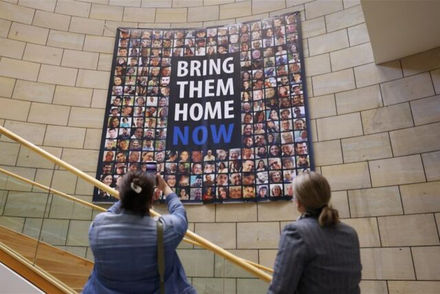 Zwei Frauen stehen vor einem Plakat mit der Aufschrift „Bring them home now“, eine fotografiert es mit ihrem Handy.
