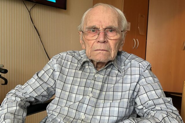 Erhard Kollitz sitzt in seinem Rollstuhl in seinem Zimmer im Altenheim. Im Hintergrund ist ein Fernseher zu sehen, ebenso ein Rollator.