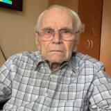 Erhard Kollitz sitzt in seinem Rollstuhl in seinem Zimmer im Altenheim. Im Hintergrund ist ein Fernseher zu sehen, ebenso ein Rollator.