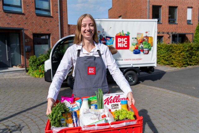 Eine junge Frau trägt eine Kiste mit Einkäufen, im Hintergrund sieht man einen Van mit Picnic-Aufschrift.