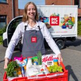 Eine junge Frau trägt eine Kiste mit Einkäufen, im Hintergrund sieht man einen Van mit Picnic-Aufschrift.