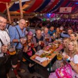 Elf Besucher in Oktoberfest-Outfits sitzen an einem länglichen Tisch, posieren für die Kamera und halten Bierkrüge hoch. Im Hintergrund sieht man eine große Menge weiterer Besucher im Festzelt.