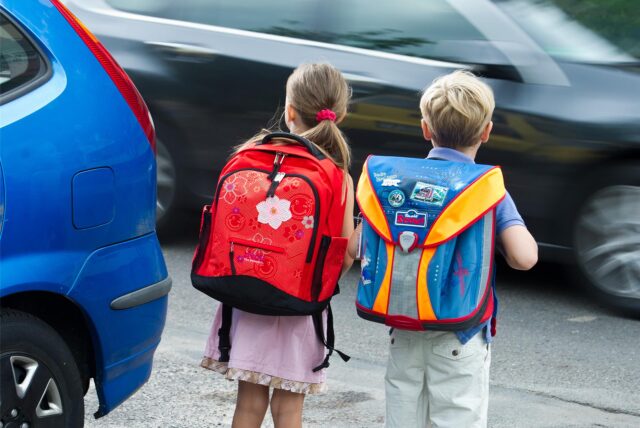Ein Junge und ein Mädchen mit Schulranzen stehen an einer befahrenen Straße mit Autos, die sie überqueren wollen.