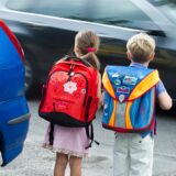 Ein Junge und ein Mädchen mit Schulranzen stehen an einer befahrenen Straße mit Autos, die sie überqueren wollen.