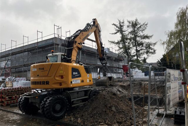 Ein großer Bagger zwischen Erdhügeln und Bauzäunen. Im Hintergrund das eingerüstete Hallenbad.