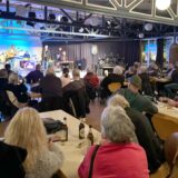 Blick in den Saal des Bürgerhauses Herten-Süd. Im Bild-Hintergrund spielt eine Musikband auf der Bühne, an Tischen davor sitzen viele Männer und Frauen.