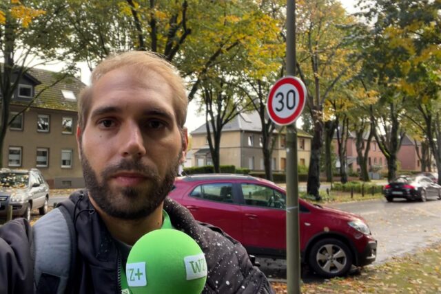 Reporter Daniel Brill steht mit einem Mikrofon in der Hand an der Waltroper Riphausstraße. Im Hintergrund parkt ein Auto neben einem Mast, an dem ein Tempo-30-Schild hängt.