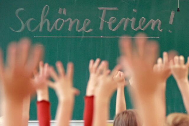 „Schöne Ferien!“ steht auf einer Tafel in einem Klassenraum, in dem die Schüler vor Freude die Hände heben.