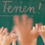 „Schöne Ferien!“ steht auf einer Tafel in einem Klassenraum, in dem die Schüler vor Freude die Hände heben.