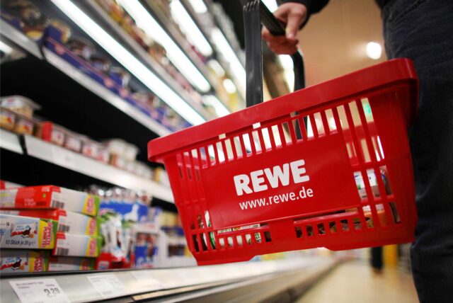 Rewe-Einkaufskorb in einer Rewe-Filiale vor einem Regal.