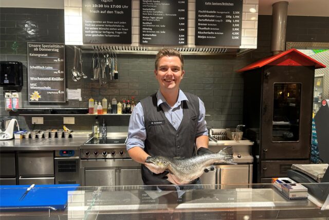 Ein Auszubildender aus Oer-Erkenschwick steht hinter der Fischtheke im Edeka Zierles und hält einen Lachs in den Händen.