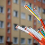 Eine Büschel Glasfaserleitungen mit ragt aus bunten Schläuchen hervor. Im Hintergrund ist ein großer Mehrfamilienhaus-Block zu sehen.