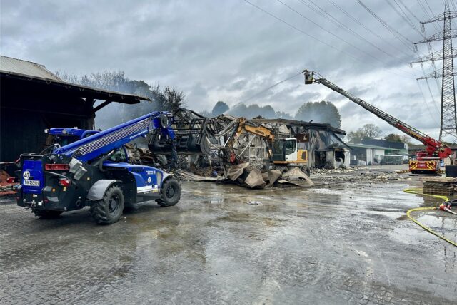 Ein Radlader des THW sowie eine Drehleiter der Feuerwehr stehen bereit, um den Brand beim Raiffeisen-Markt in Hünxe unter Kontrolle zu bringen.