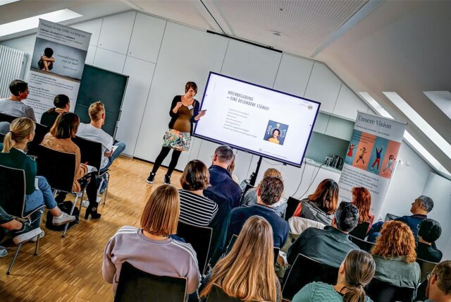 Dr. Stefanie Marzian vom Verein Herausforderung steht beim Info-Tag „Wellenbrecher“ vor einem Raum voller Menschen und hält einen Vortrag.