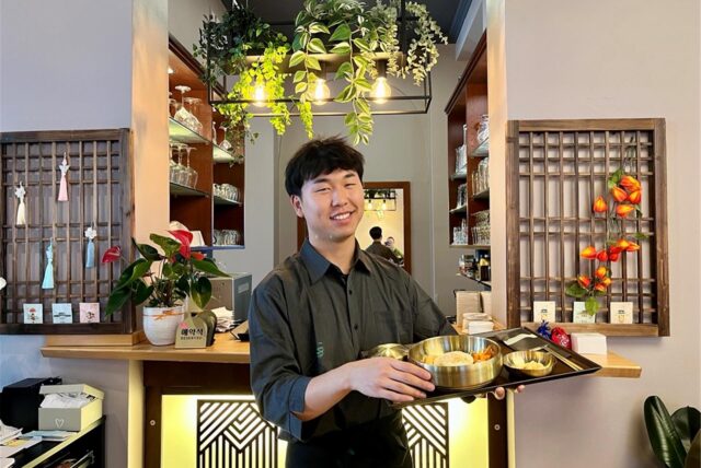 Yejun Park steht im The Korean in Dortmund und hält ein Tablett mit Speisen in den Händen.