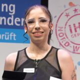 Jochen Grütters, Hannah Bafs und Melanie Baum stehen nebeneinander. Hannah Bafs trägt eine kleine Glas-Trophäe, Melanie Baum präsentiert Bafs‘ Urkunde.