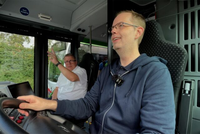 Ein Mann sitzt am Steuer eines Busses, dahinter sitzt ein Mann und erklärt etwas.