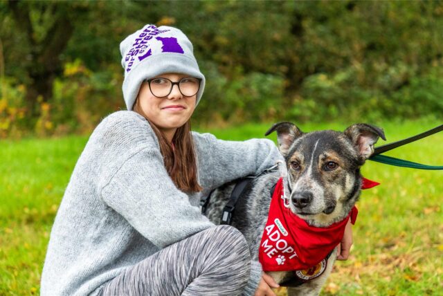 Josefin Weber kniet neben Rüde Karlchen, der ein rotes Halstuch mit der Aufschrift „Adopt Me“ trägt.
