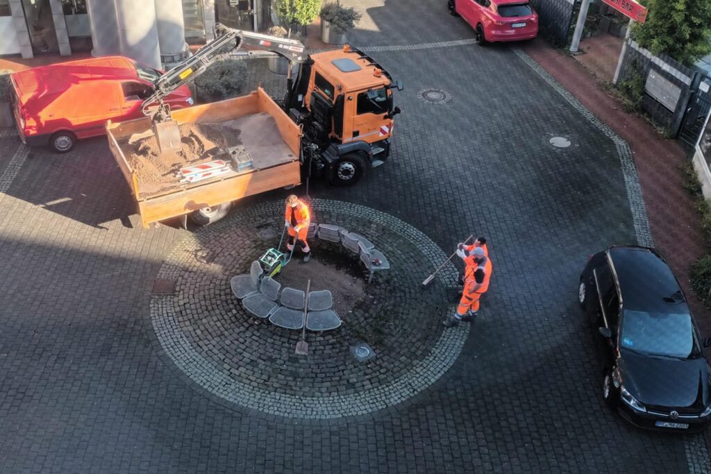 Eine Baumscheibe auf dem Platz an der Turmstraße und Wiethofstraße in der Innenstadt von Recklinghausen wird von einem Arbeiter in orangefarbener Kleidung mit einer Rüttelplatte verdichtet. Eine Baumscheibe auf dem Platz an der Turmstraße und Wiethofstraße in der Innenstadt von Recklinghausen wird von einem Arbeiter in orangefarbener Kleidung mit einer Rüttelplatte verdichtet.