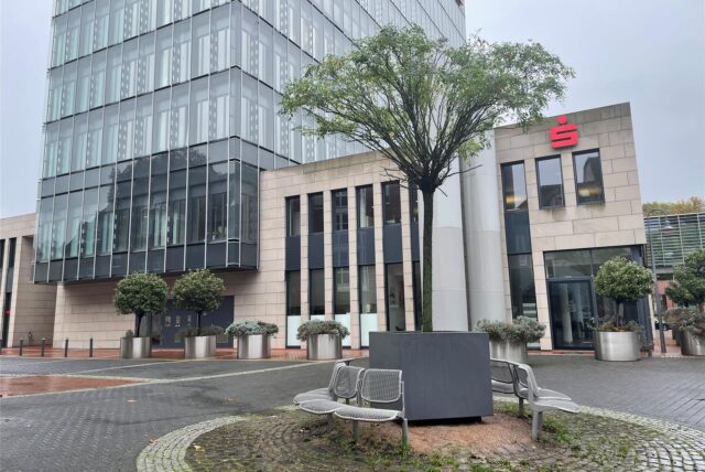 Ein Baum in einem quadratischen Topf steht auf einer Baumscheibe auf dem Platz hinter der Sparkasse am Herzogswall in Recklinghausen.