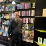 Wolfgang Tänzer steht im Bücherwurm vor Bücherregalen und hält ein Buch mit mehrfarbigem Farbschnitt in der Hand.