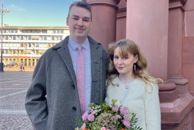 Das junge Ehepaar Glen und Vivian steht, nach ihrer Standesamtlichen Trauung auf dem Rathausplatz in Dortmund. Im Hintergrund sieht man die Säulen des Standesamts.