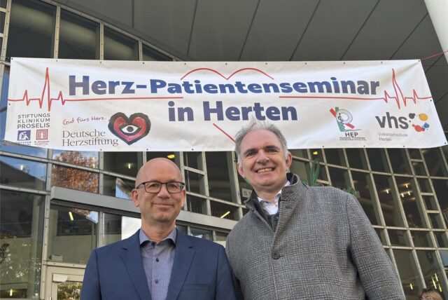 Horst Neubauer und Klaus Hillebrand stehen vor dem Eingang des Glashauses in der Hertener Innenstadt. Hinter ihnen hängt ein großes Banner mit der Aufschrift Her-Patientenseminar in Herten.