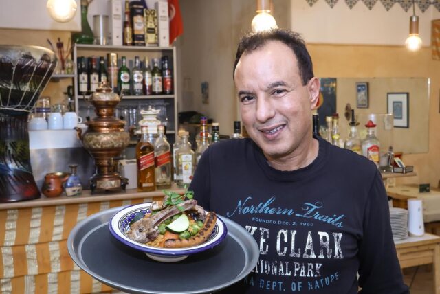 Kamel Zouaghi, Koch und Inhaber vom Restaurant Meridien in Dortmund. Er hält einen Teller mit einem Hauptgericht in der Hand, im Hintergrund erkennt man den Thekenbereich des Restaurants.