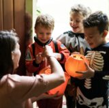 Mit dem richtigen Halloween-Spruch bekommen diese drei verkleideten Kinder von einer Frau Süßigkeiten überreicht.