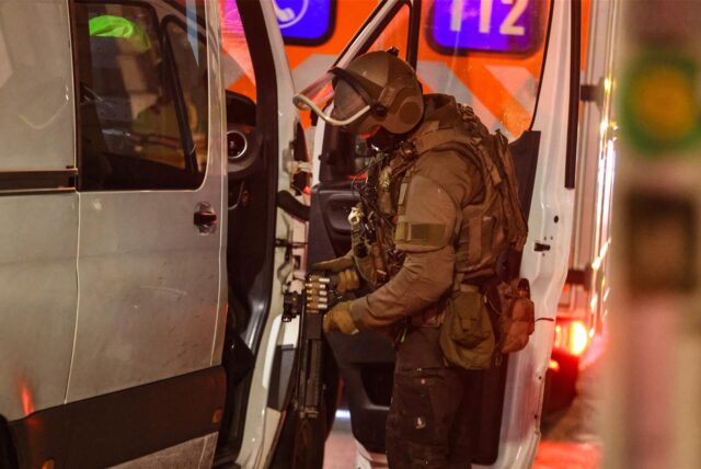 Eine Person in SEK Kleidung steht an der Tür eines silbernen Vans und hantiert an einer Waffe.
